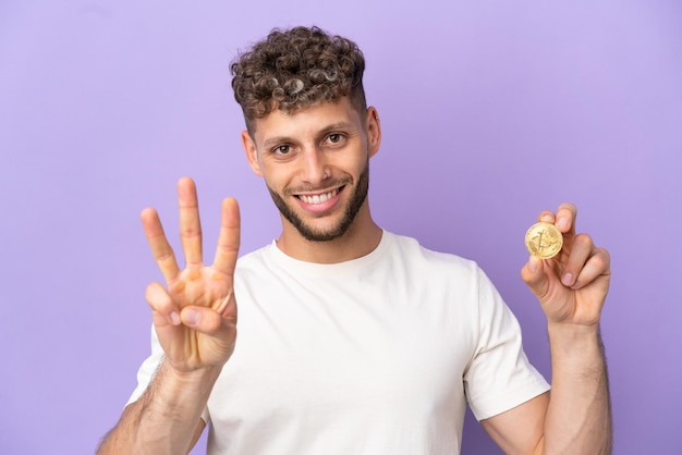 Joven caucásico sosteniendo un Bitcoin aislado de fondo morado feliz y contando tres con los dedos