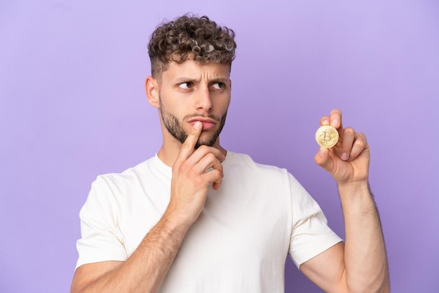 Joven caucásico sosteniendo un Bitcoin aislado de fondo morado que tiene dudas mientras mira hacia arriba