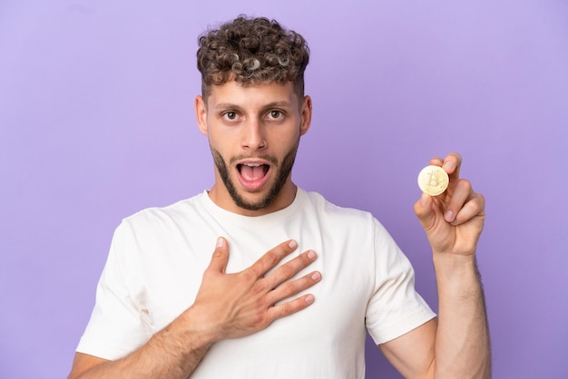 Joven caucásico sosteniendo un Bitcoin aislado en un fondo morado sorprendido y conmocionado mientras miraba a la derecha