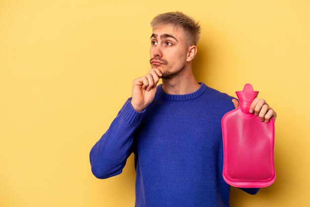 Joven caucásico sosteniendo una bolsa de agua aislada en un fondo amarillo mirando de lado con expresión dudosa y escéptica.