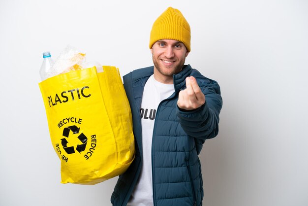 Joven caucásico sosteniendo una bolsa llena de botellas de plástico para reciclar aislado en fondo blanco haciendo gesto de dinero