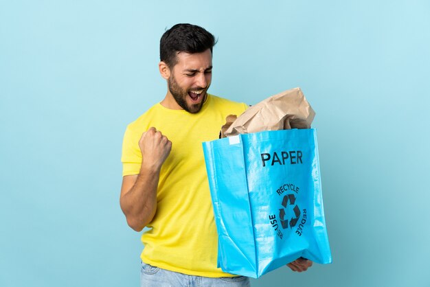 Joven caucásico sosteniendo una bolsa de reciclaje aislada sobre fondo azul celebrando una victoria