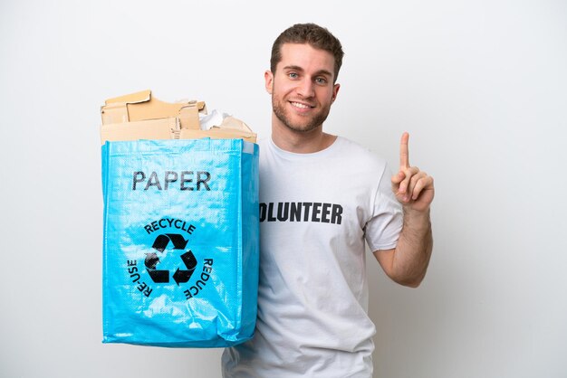 Joven caucásico sosteniendo una bolsa de reciclaje llena de papel para reciclar aislado sobre fondo blanco apuntando hacia una gran idea