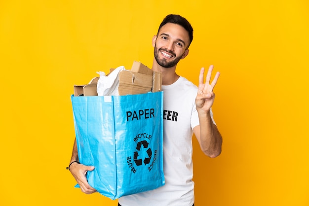 Joven caucásico sosteniendo una bolsa de reciclaje llena de papel para reciclar aislado sobre fondo blanco feliz y contando tres con los dedos