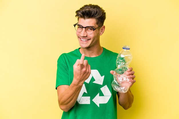 Foto joven caucásico sosteniendo una botella de plástico para reciclar aislado en un fondo amarillo apuntándote con el dedo como si invitaras a acercarte