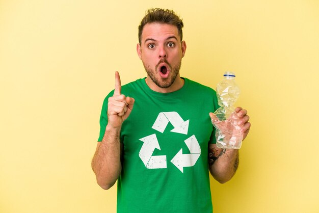 Joven caucásico sosteniendo una botella de plástico para reciclar aislado en un fondo amarillo con una gran idea, concepto de creatividad.