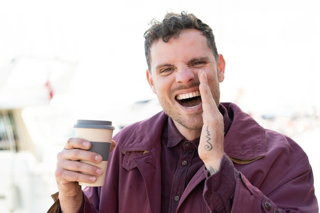 Joven caucásico sosteniendo un café para llevar al aire libre gritando con la boca abierta