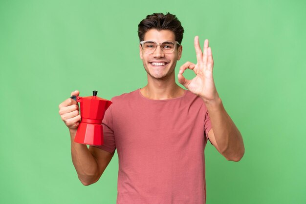 Joven caucásico sosteniendo una cafetera sobre un fondo aislado que muestra el signo de ok con los dedos