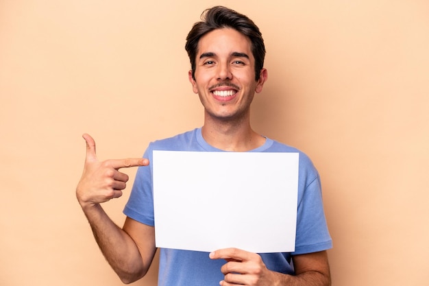 Joven caucásico sosteniendo un cartel aislado en una persona de fondo beige señalando con la mano un espacio de copia de camisa orgulloso y confiado