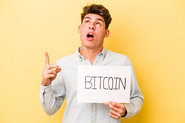 Joven caucásico sosteniendo un cartel de bitcoin aislado en un fondo amarillo apuntando hacia arriba con la boca abierta