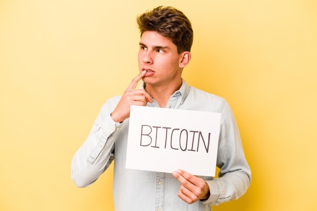 Joven caucásico sosteniendo un cartel de bitcoin aislado en un fondo amarillo relajado pensando en algo mirando un espacio de copia