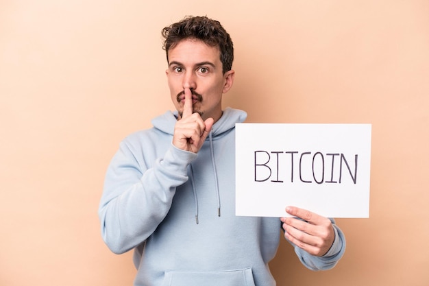 Joven caucásico sosteniendo un cartel de bitcoin aislado de fondo beige guardando un secreto o pidiendo silencio