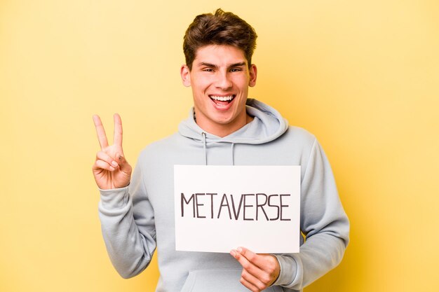 Joven caucásico sosteniendo un cartel de metaverso aislado en un fondo amarillo alegre y despreocupado mostrando un símbolo de paz con los dedos