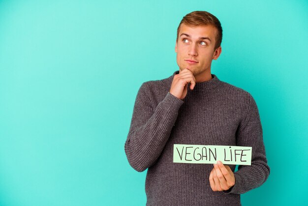 Joven caucásico sosteniendo un cartel de vida vegana aislado sobre fondo azul mirando hacia los lados con expresión dudosa y escéptica.