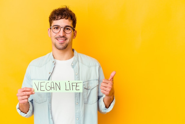 Joven caucásico sosteniendo un cartel de vida vegana aislado sonriendo y levantando el pulgar hacia arriba