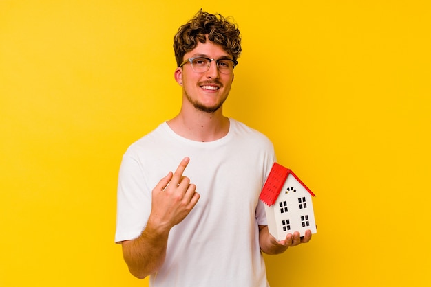 Joven caucásico sosteniendo una casa modelo aislada sobre fondo amarillo apuntando con el dedo hacia usted como si estuviera invitando a acercarse.