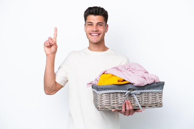 Joven caucásico sosteniendo una cesta de ropa aislada de fondo blanco mostrando y levantando un dedo en señal de lo mejor