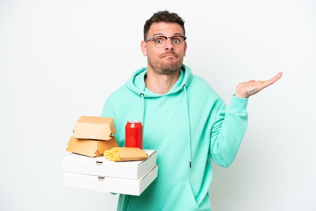 Joven caucásico sosteniendo comida rápida aislado de fondo blanco que tiene dudas mientras levanta las manos