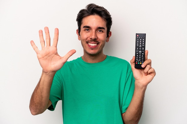 Joven caucásico sosteniendo un control remoto de tv aislado en fondo blanco sonriendo alegre mostrando el número cinco con los dedos.