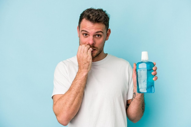 Joven caucásico sosteniendo enjuague bucal aislado de fondo azul mordiéndose las uñas nervioso y muy ansioso