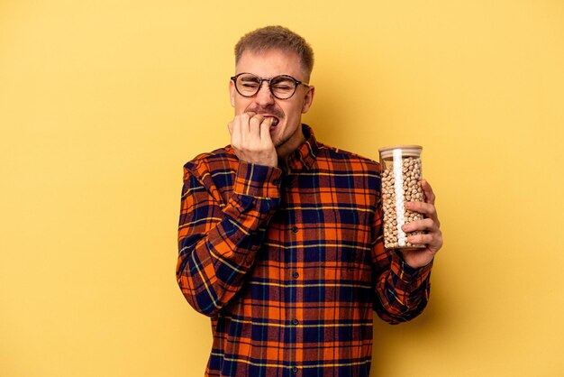 Joven caucásico sosteniendo garbanzos aislados en un fondo amarillo mordiéndose las uñas, nervioso y muy ansioso.