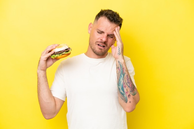 Joven caucásico sosteniendo una hamburguesa aislada de fondo amarillo con dolor de cabeza