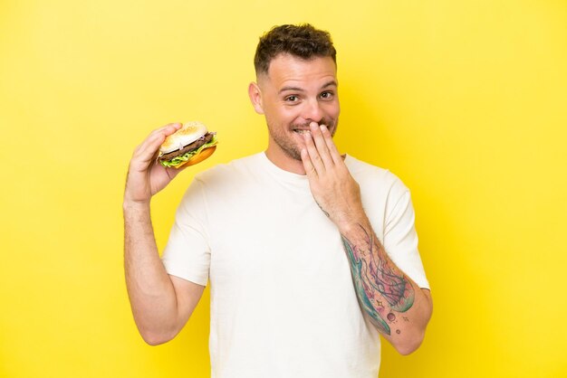 Joven caucásico sosteniendo una hamburguesa aislada de fondo amarillo feliz y sonriente cubriendo la boca con la mano