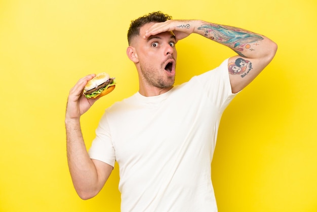 Joven caucásico sosteniendo una hamburguesa aislada de fondo amarillo haciendo un gesto sorpresa mientras mira hacia un lado