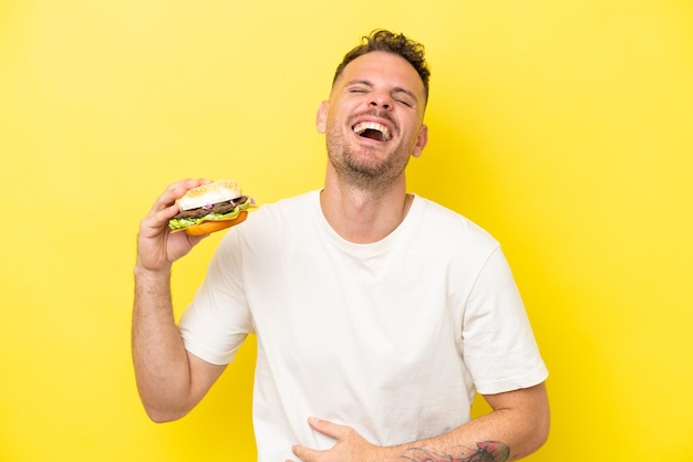 Joven caucásico sosteniendo una hamburguesa aislada de fondo amarillo sonriendo mucho