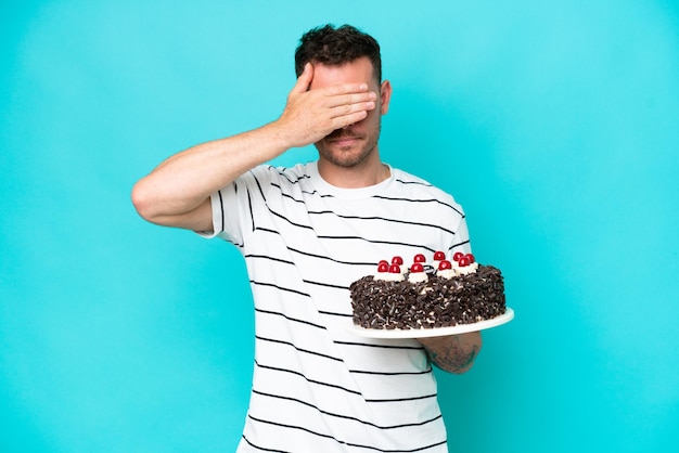 Joven caucásico sosteniendo pastel de cumpleaños aislado de fondo azul cubriendo los ojos con las manos No quiero ver algo