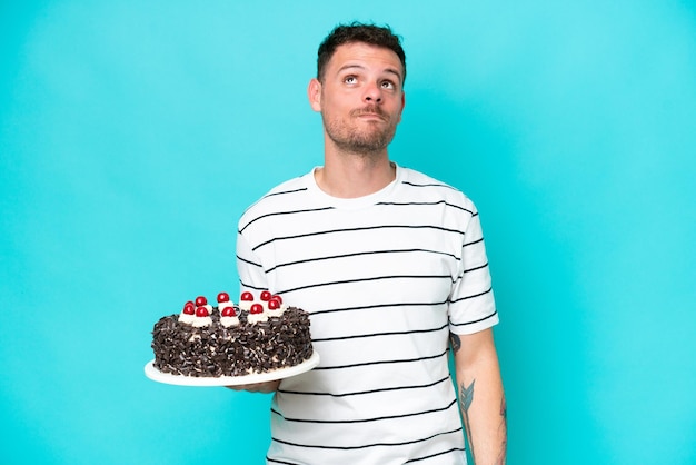 Foto joven caucásico sosteniendo pastel de cumpleaños aislado de fondo azul y mirando hacia arriba