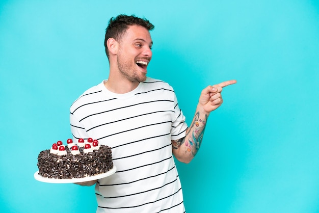 Foto joven caucásico sosteniendo pastel de cumpleaños aislado de fondo azul señalando con el dedo hacia un lado y presentando un producto