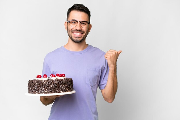 Joven caucásico sosteniendo pastel de cumpleaños aislado de fondo blanco apuntando hacia un lado para presentar un producto