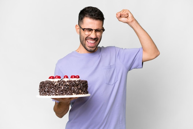 Joven caucásico sosteniendo pastel de cumpleaños aislado de fondo blanco celebrando una victoria