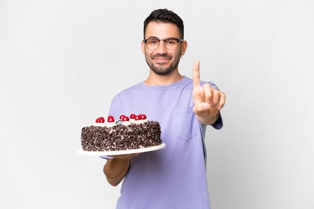 Joven caucásico sosteniendo pastel de cumpleaños aislado de fondo blanco mostrando y levantando un dedo