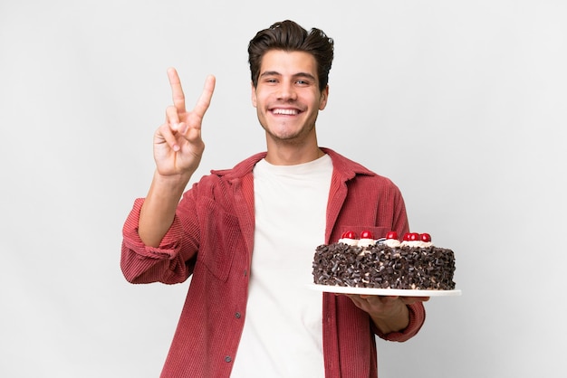 Joven caucásico sosteniendo pastel de cumpleaños sobre un fondo aislado sonriendo y mostrando el signo de la victoria