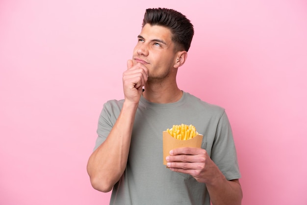 Joven caucásico sosteniendo patatas fritas aisladas de fondo rosa con dudas