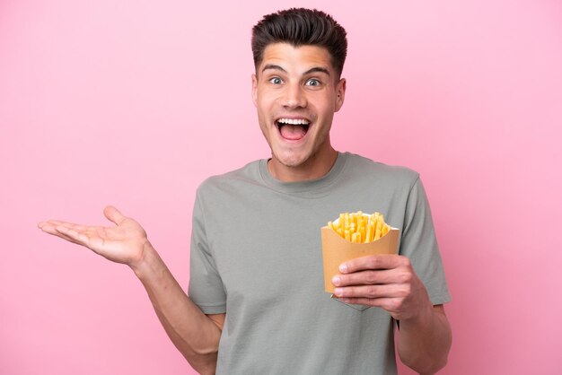 Joven caucásico sosteniendo patatas fritas aisladas de fondo rosa con expresión facial conmocionada