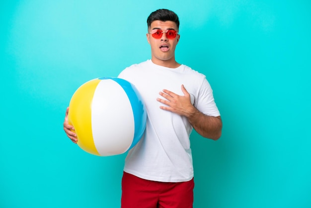 Joven caucásico sosteniendo una pelota de playa aislada de fondo azul sorprendido y conmocionado mientras miraba a la derecha