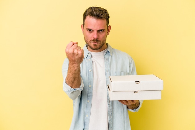 Joven caucásico sosteniendo pizzas aisladas de fondo amarillo mostrando el puño a la cámara con una expresión facial agresiva