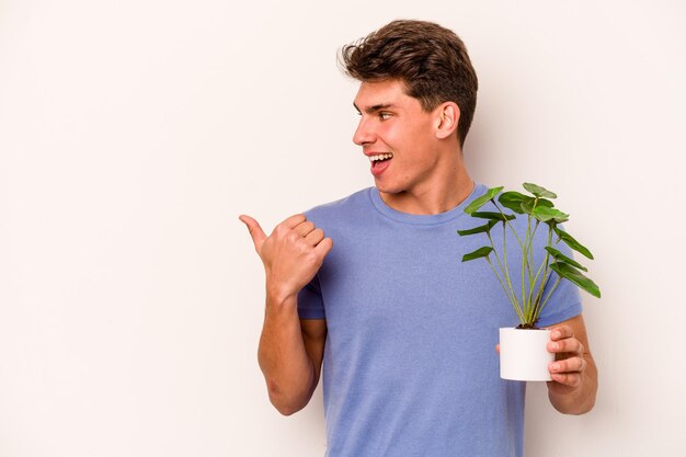 Joven caucásico sosteniendo una planta aislada en los puntos de fondo blanco con el dedo pulgar lejos riendo y despreocupado