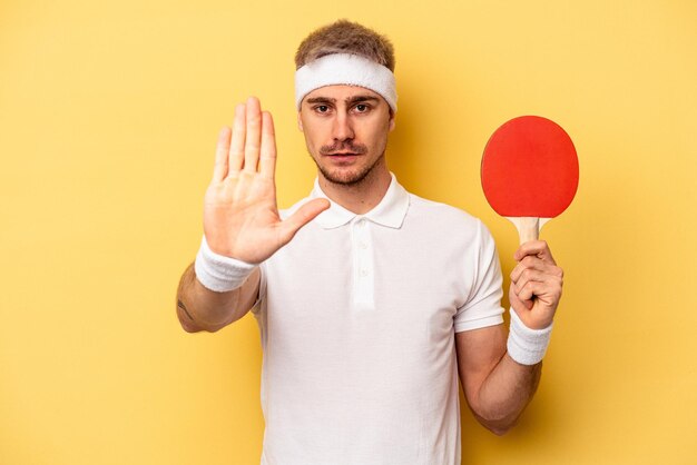 Joven caucásico sosteniendo raquetas de ping pong aisladas en un fondo amarillo de pie con la mano extendida mostrando la señal de alto, impidiéndote.