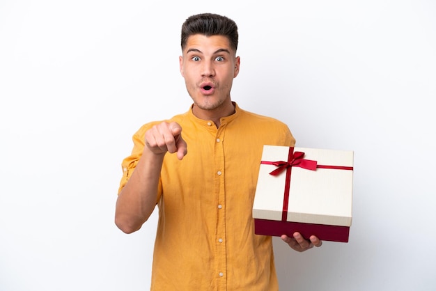 Joven caucásico sosteniendo un regalo aislado de fondo blanco sorprendido y apuntando al frente