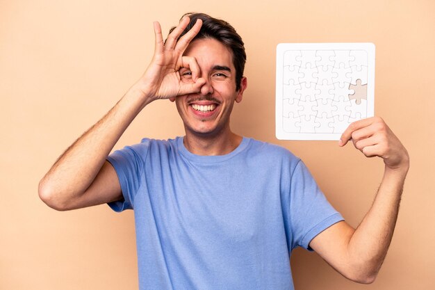 Joven caucásico sosteniendo un rompecabezas aislado en un fondo beige emocionado manteniendo el gesto correcto en el ojo.