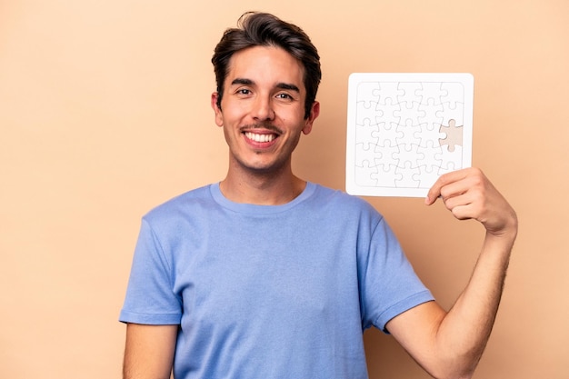 Joven caucásico sosteniendo un rompecabezas aislado en un fondo beige feliz, sonriente y alegre.