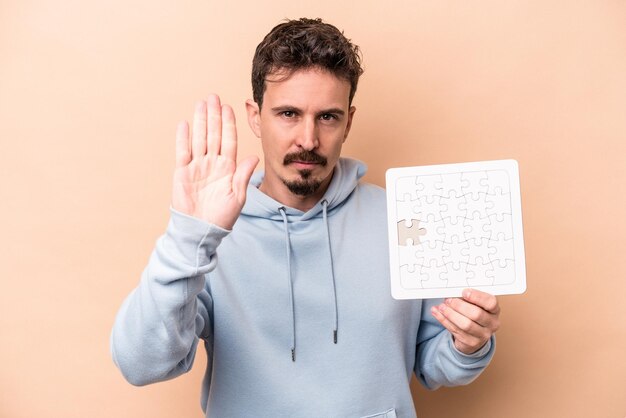 Joven caucásico sosteniendo un rompecabezas aislado en un fondo beige de pie con la mano extendida mostrando la señal de alto, impidiéndote.