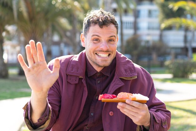 Foto joven caucásico sosteniendo sashimi al aire libre saludando con la mano con expresión feliz