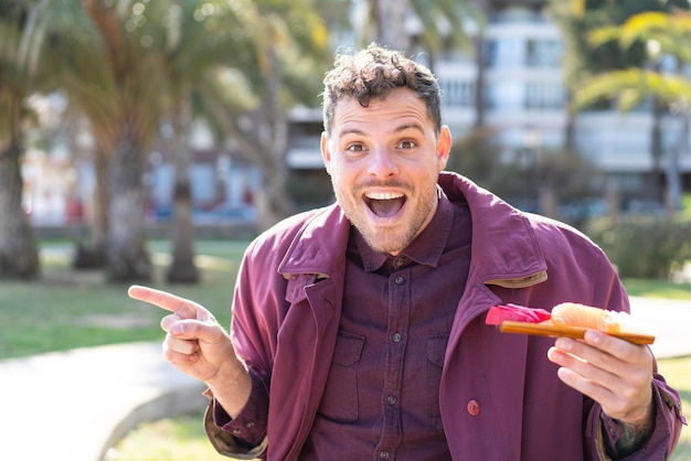 Joven caucásico sosteniendo sashimi al aire libre sorprendido y señalando con el dedo hacia un lado