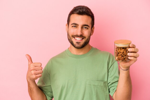 Joven caucásico sosteniendo un tarro de almendras aislado en la pared rosa sonriendo y levantando el pulgar hacia arriba