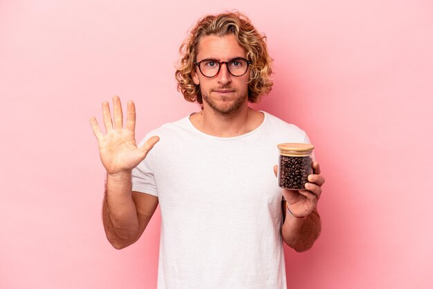 Joven caucásico sosteniendo un tarro de café aislado en un fondo rosa sonriendo alegre mostrando el número cinco con los dedos.